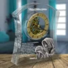 Transparent curved glass sculpture enclosing a gold clock face with Roman numerals, overlaid with white Slovak text; a kneeling figure is visible nearby on a wooden floor.