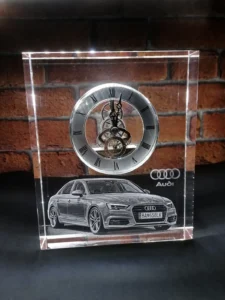 ChatGPT Image 11 9 2026 20_36_18 | Gravírovanie fotiek do skla - darček s venovaním Clear acrylic desk ornament with a gear-driven clock above a grayscale Audi car image and logo, set against a brick wall.