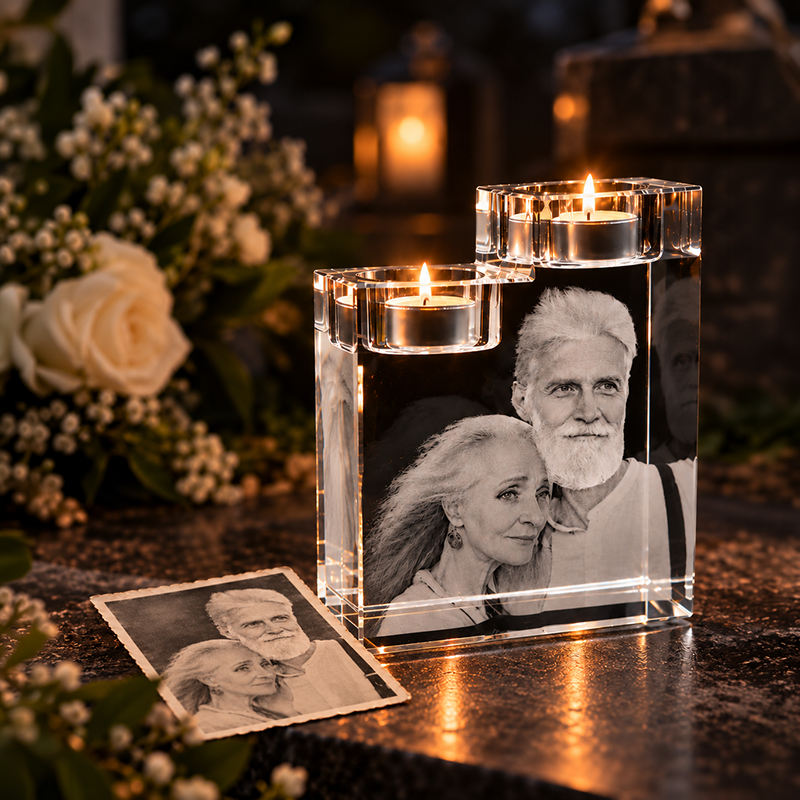 Crystal memorial block with etched portrait of an elderly couple and two lit tealight candles on top, in a somber setting with flowers nearby.